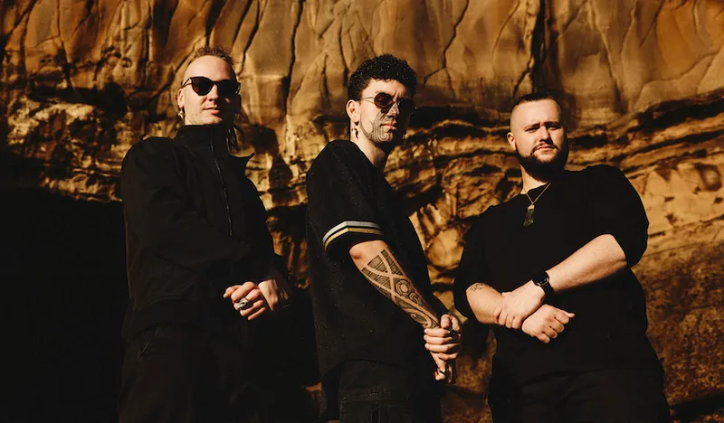 The members of Alien Weaponry stand in black against a brown cliff face
