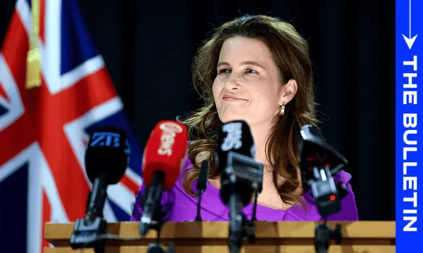 Nicola Willis stands at a podium speaking into several microphones. She wears a purple outfit and smiles slightly. Behind her, a flag with red, white, and blue colors is partially visible, suggesting an official event or announcement.