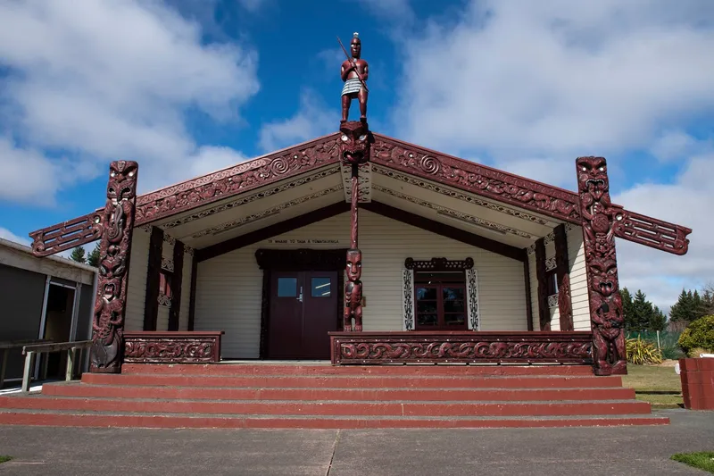 A traditional Māori meeting house with intricate red carvings and a statue on top. The building has a peaked roof with ornamental details and sits on a raised platform with steps. The sky is partly cloudy.