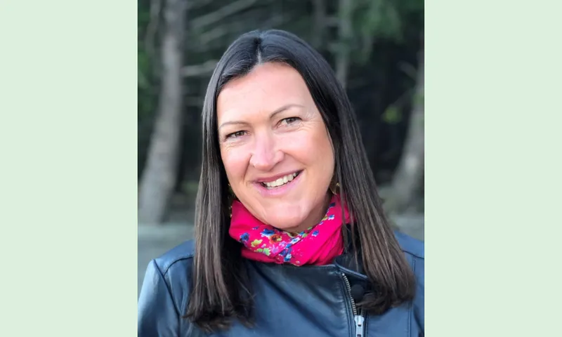 a white whoman with dark hair wearing a pink scarf with trees in the background smiling slightly