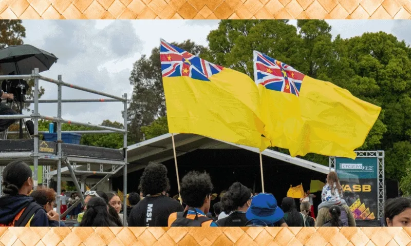A public event with people gathered in front of a stage. Two large yellow flags with red and blue patterned corners are prominently displayed. Trees and a scaffold are visible in the background.