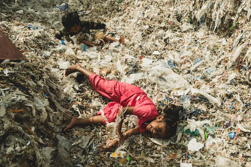 Two children play joyfully in a large pile of plastic waste. The child in front, wearing a pink outfit, has an expression of delight. The other child in dark clothing lies further back. The scene highlights a stark contrast between play and pollution.