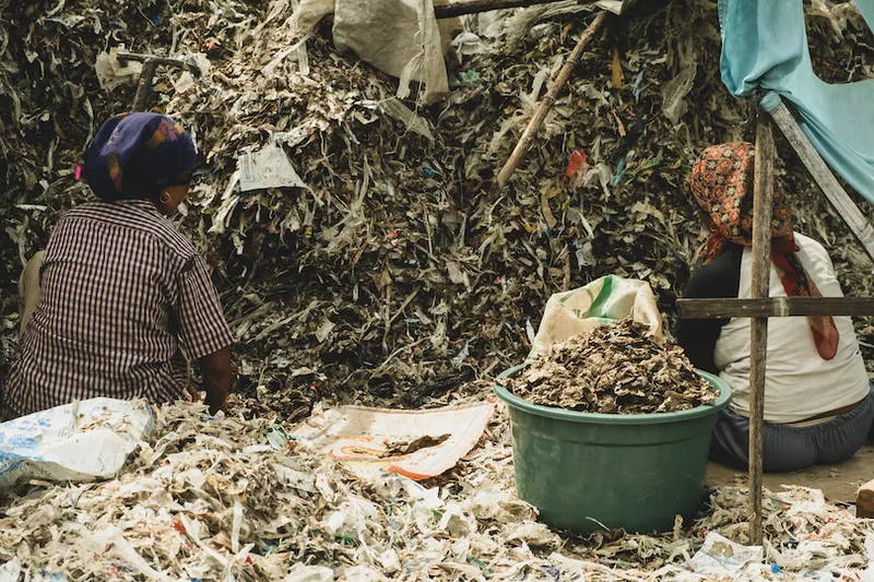 Two people work amidst large piles of shredded material and debris. One sits beside a tub filled with smaller shredded pieces. They appear focused on sorting or processing the materials. Fabric covers part of the piles, and a makeshift shelter is visible.