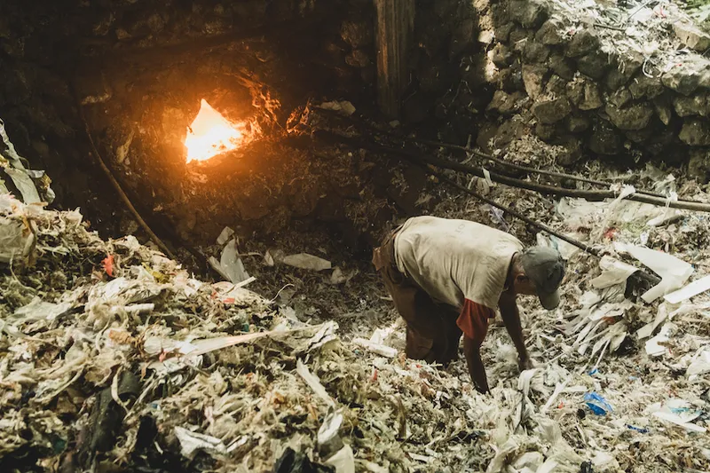 A person bends over, sorting through piles of trash near a large open fire in an industrial setting. The stone walls and scattered debris indicate a challenging work environment.