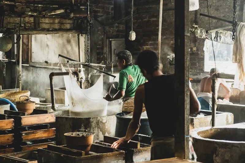 Workers in a rustic workshop setting, engaged in processing materials using traditional methods. The room is filled with wooden equipment and steam, creating a warm, industrious atmosphere.
