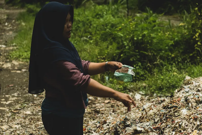 A person wearing a dark headscarf stands by a large pile of trash, holding a mask in one hand, in an outdoor setting surrounded by greenery. The scene conveys an environmental concern.