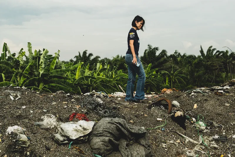 A person walks on a mound of dirt and scattered trash, holding a white object. They are surrounded by green banana trees under a cloudy sky. The scene suggests a focus on environmental pollution or waste management.