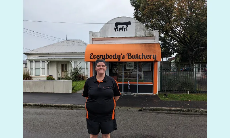 LIsa willert, a broad white woman wearing a black tshirt and shorts, stands in front of Everybody's butchery, a shop with an orange sign