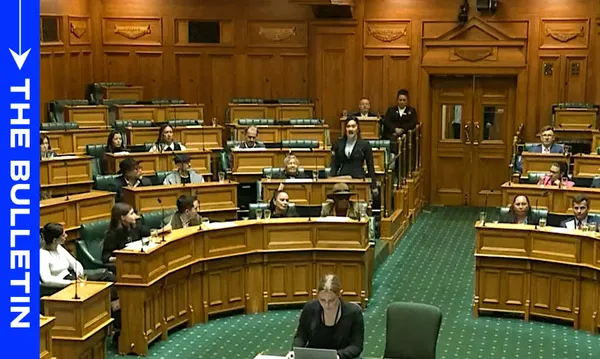 People are seated in a parliamentary chamber with wooden interiors and green seats. A person is standing and speaking, while others listen. A large screen displays the text "The Bulletin" on the left side.