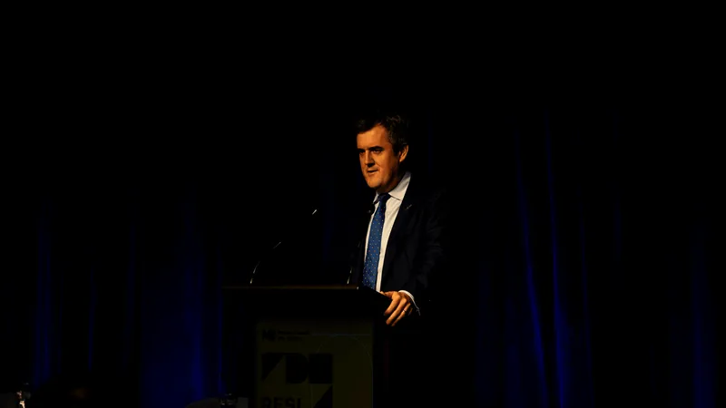A man in a suit speaks at a podium on a dimly lit stage, with a dark blue curtain backdrop.