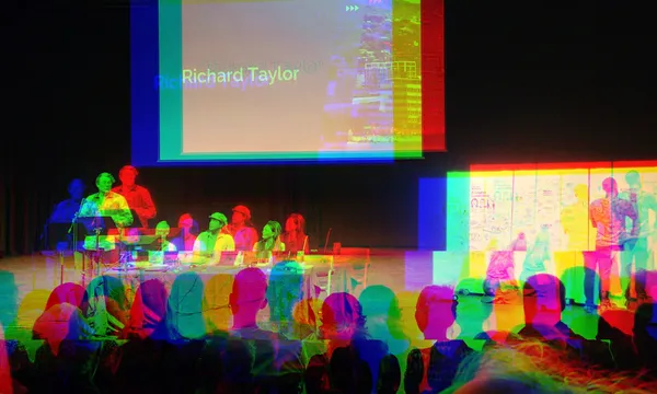 Audience watching a colorful, glitch-effect presentation in a dimly lit room. A group of people is seated at desks on stage, with a large screen displaying text, including the name "Richard Taylor.