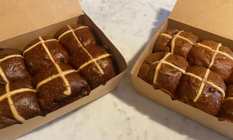 Two open cardboard boxes on a marble surface, each containing six dark brown hot cross buns with white icing crosses on top.