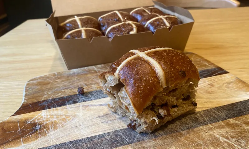 An open cardboard box on a wooden table, containing six dark brown hot cross buns with white icing crosses on top, and a single bun on a wooden board