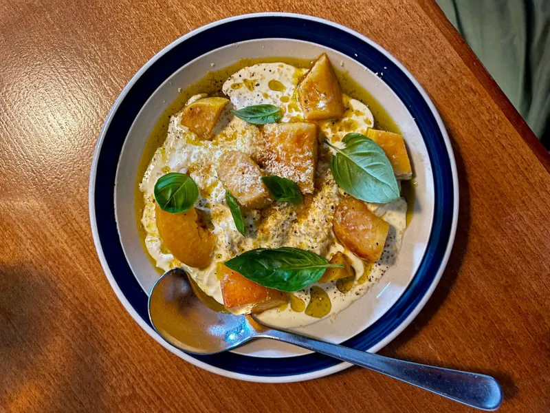 A bowl of creamy stracciatella cheese topped with pieces of peach, basil leaves, and grated cheese. A silver spoon rests on the bowl's edge, which is on a wooden table. Drizzles of olive oil and pepper are visible.
