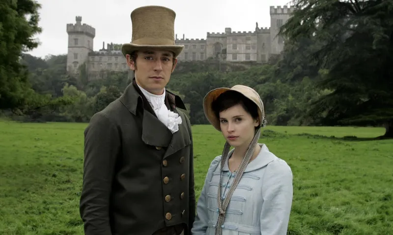 Felicity Jones as Catherine Morland and JJ Feild as Henry Tilney in the 2007 film of Northanger Abbey. They are standing in costume on a grassy field in front of a huge Abbey.