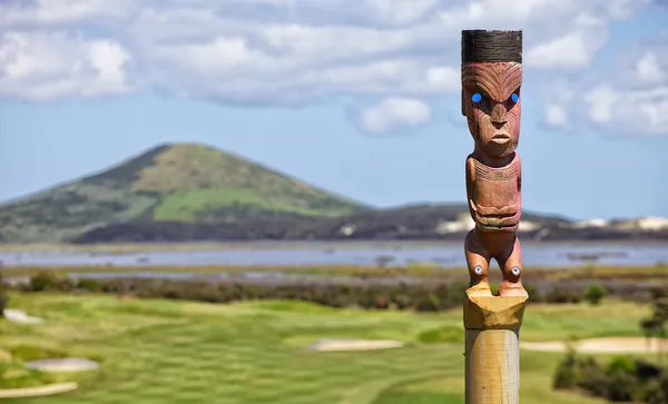 A wooden pou whenua with blue eyes is mounted on a post, overlooking a green golf course with a mountain and cloudy sky in the background.