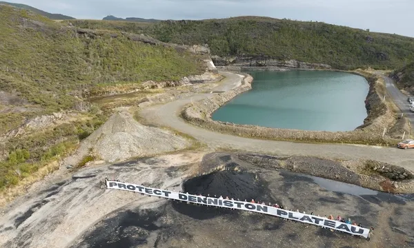 a lake and a shallow pit of dark earth surrounded by bouncy green bush, with a line of people holding a banner that reads "protect denniston plateau"