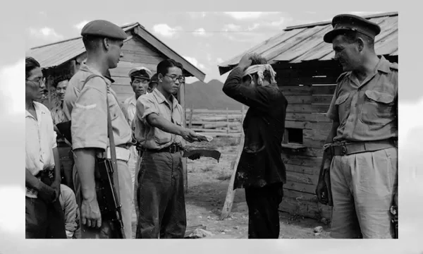 A group of uniformed and plainclothes men surround a person with hands on their head, standing near wooden huts. One man holds a pile of banknotes while others watch. A New Zealand soldier with a rifle stands nearby.