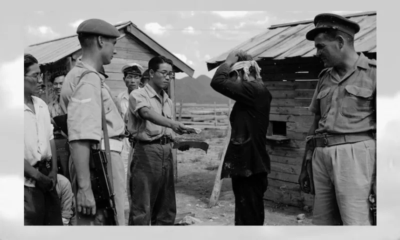 A group of uniformed and plainclothes men surround a person with hands on their head, standing near wooden huts. One man holds a pile of banknotes while others watch. A New Zealand soldier with a rifle stands nearby.