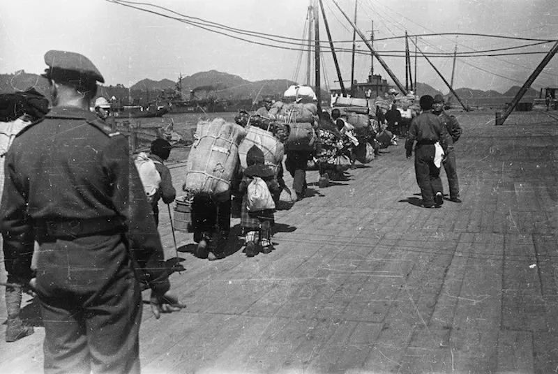 A group of people carrying large bundles and bags walk in a line along a dock, watched by uniformed soldiers. A ship and mountains are visible in the background. The image is in black and white.