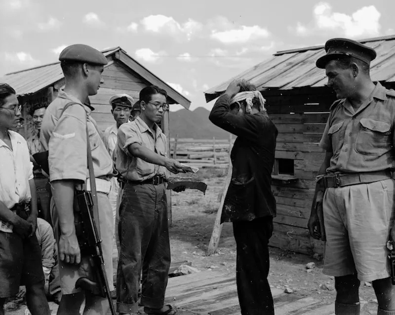 A group of uniformed and plainclothes men surround a person with hands on their head, standing near wooden huts. One man holds a pile of banknotes while others watch. A New Zealand soldier with a rifle stands nearby.