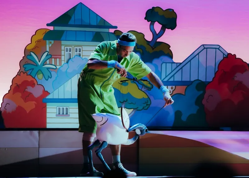 A performer in green clothes manipulates a white bird puppet on stage, with a colourful, cartoon-like backdrop of houses and trees behind him.