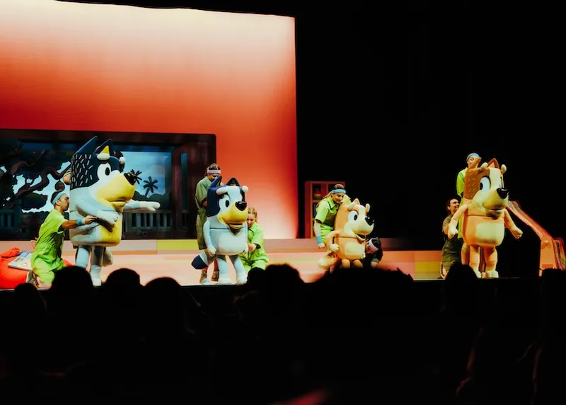 People in green outfits control large dog character costumes on stage in front of an orange-lit backdrop, performing for an audience in a theatre setting.