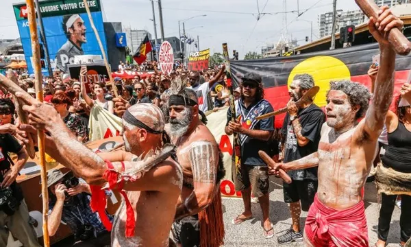 A large group of people, some with painted faces and bodies, participate in a protest carrying Aboriginal flags and banners. Several individuals play wooden instruments in the foreground.