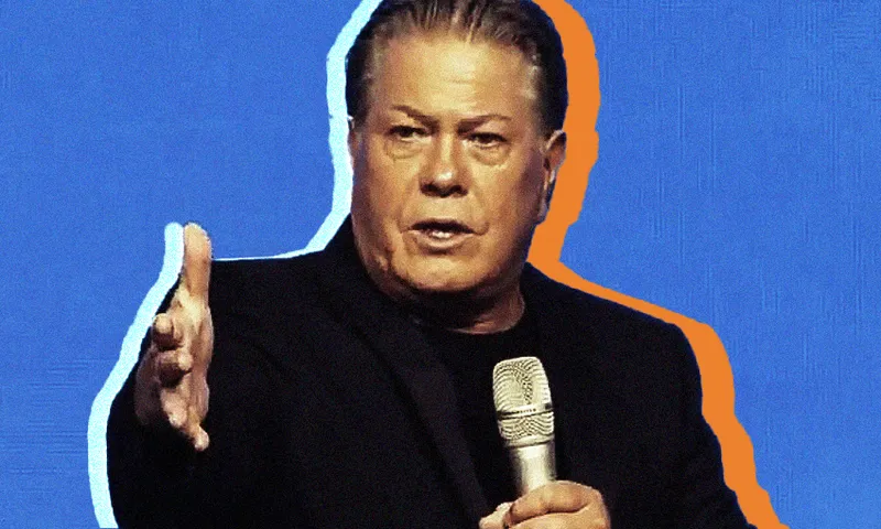 A man in a black suit holds a microphone and gestures with his other hand against a blue background outlined in orange.