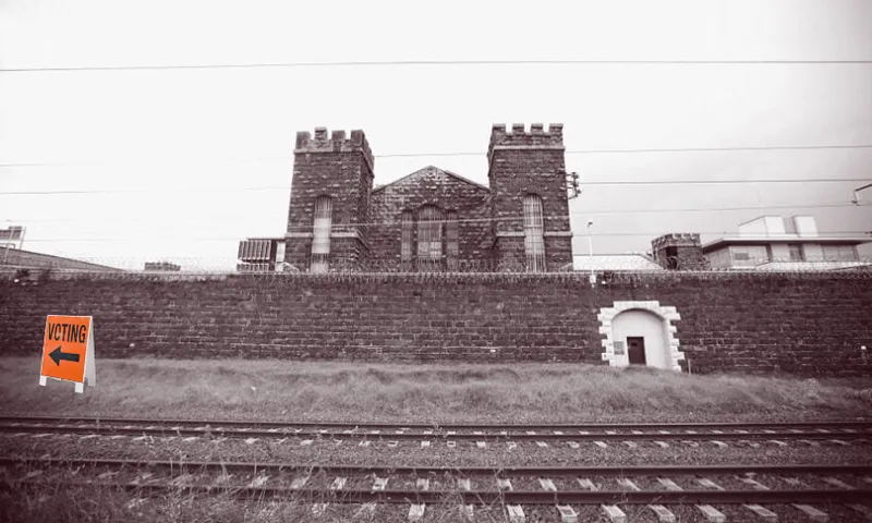 A large stone prison building stands behind a high wall and railway tracks. A bright orange sign on the grass reads "VOTING" with an arrow pointing left.