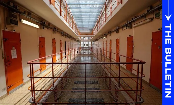 A hallway in a prison with orange cell doors on both sides, a metal walkway and railings, and a glass ceiling. A blue vertical banner on the right reads "THE BULLETIN.