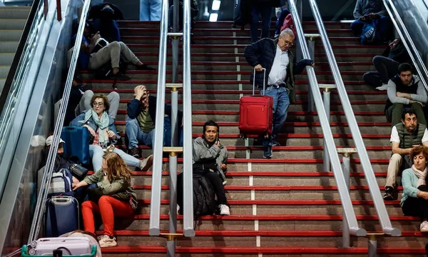 People sit with luggage on red-carpeted stairs between two escalators in a busy public space, while a man with a red suitcase walks down the steps.