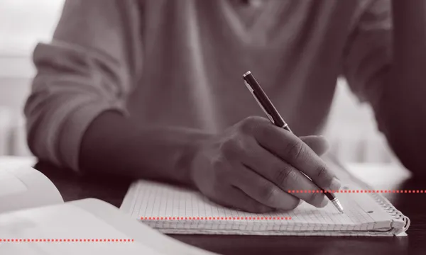 A greyscale photograph of a student writing with a pen, on lined paper.
