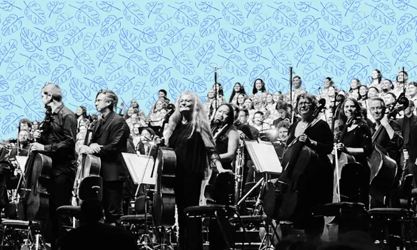 A black-and-white photo of an orchestra with musicians standing and holding their instruments, with a large choir behind them and a blue background decorated with leaf patterns.