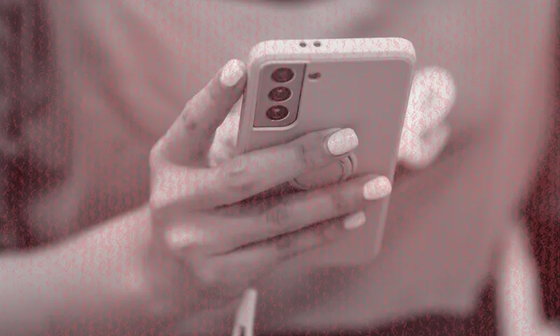 A person holding a smartphone with a white case, using their thumb to interact with the screen. The image has a red filter and shows only their hands and part of their torso.