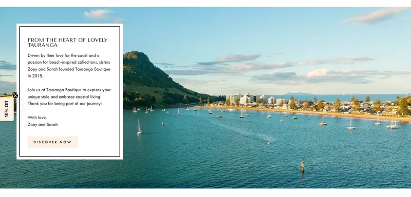 A scenic coastal view of Tauranga with calm blue waters, sailboats, a green mountain in the background, and waterfront buildings; an overlay text box shares the story of Tauranga Boutique’s founders, Zoey and Sarah.