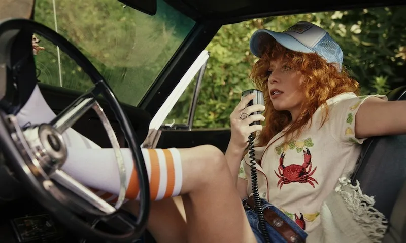 A woman with red curly hair sits inside a car, with her feet on the steering wheel. She is talking into a hand-held radio receiver and wearing a blue cap. She has knee-high white socks with an orange stripe on them.