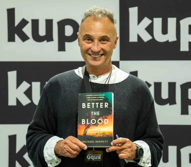 Photograph of Māori man grinning, holding up a copy of his book Better the Blood.