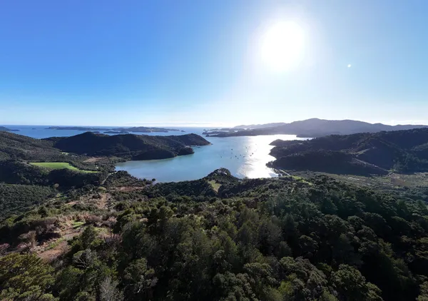 A scenic view of a bay surrounded by forested hills, with small islands in the distance and the sun shining brightly over a calm, blue sea.