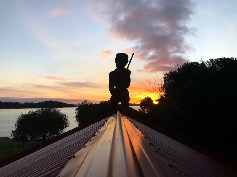 A silhouetted tekateka stands on the prow of a marae at sunrise, with a glowing sky, calm water and dark trees in the background.