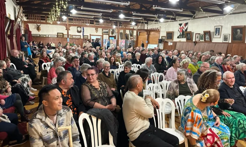 A photograph of a crowd of people seated on white chairs in an old fashioned village hall.