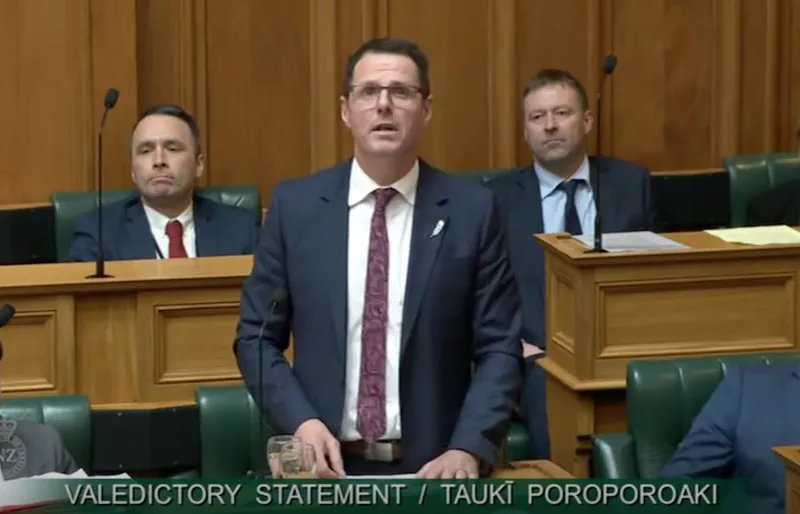 A man in a suit stands and speaks at a podium in a formal chamber, with three men seated behind him. Text at the bottom reads, "VALEDICTORY STATEMENT / TAUKI POROPOROAKI.