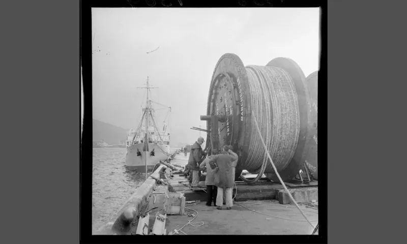 a black and white photo of big reels with a cable and a sea and boat in the background
