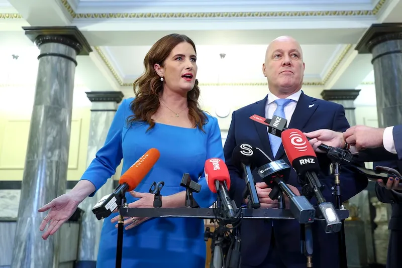 A woman in a blue dress and a man in a blue suit stand side by side speaking at a press conference, surrounded by multiple microphones and reporters in a marble-floored building.