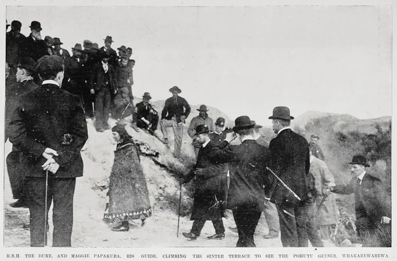 A group of men in formal clothing, some with canes and hats, stand and watch as a woman in traditional dress guides another man up a rocky hillside. Text below identifies the guide as Maggie Papakura.