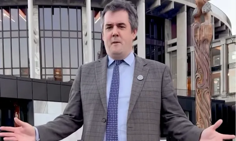 A man (Chris Bishop) in a gray suit and tie stands outdoors with arms open in front of a modern building with large windows and a tall carved wooden post.