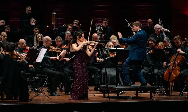 a violinist in a sparkling dress plays while a conductor in a blue shirt is in a crouching position, almost looking like he's dancing, as musicians play behind him. (mostly the violiins but the top of a harp is poking out too)