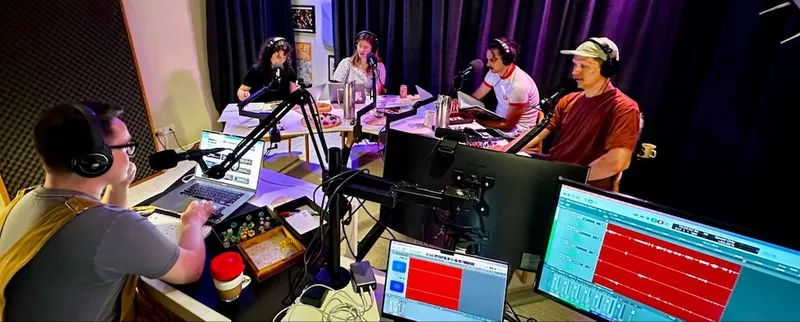 five people sit around a large table with headphones on, speaking into microphones