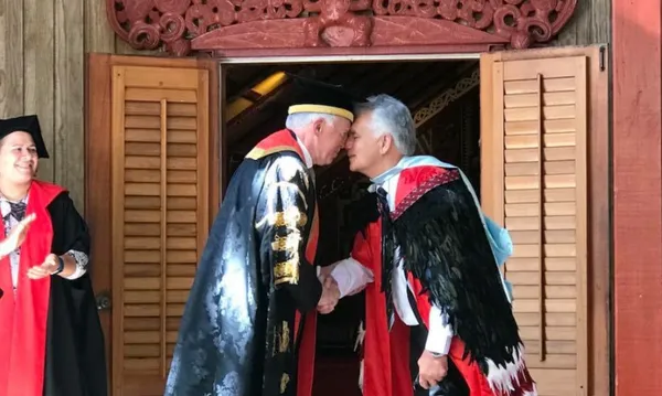 Two men in ceremonial academic dress greet each other with a hongi (traditional Māori greeting) outside a carved wharenui (meeting house). One wears a university chancellor’s gown, and the other a korowai (ceremonial cloak), symbolising the meeting of Māori and Western academic traditions.