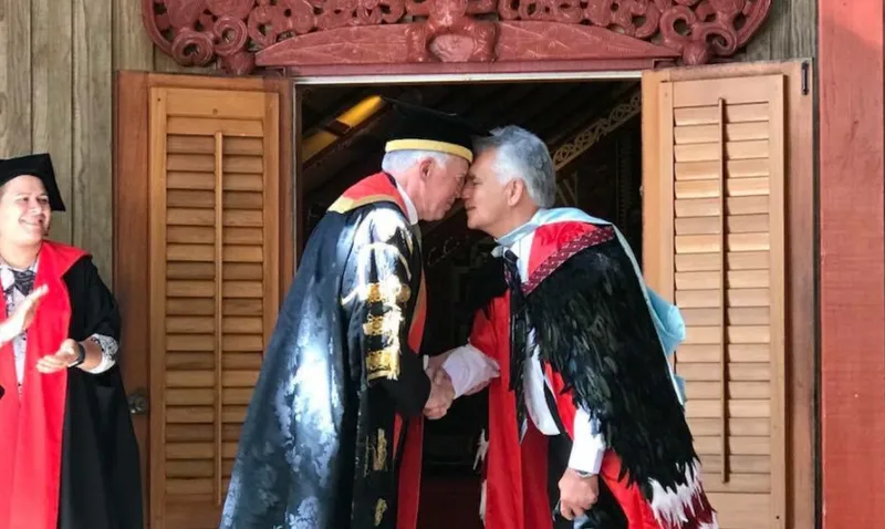 Two men in ceremonial academic dress greet each other with a hongi (traditional Māori greeting) outside a carved wharenui (meeting house). One wears a university chancellor’s gown, and the other a korowai (ceremonial cloak), symbolising the meeting of Māori and Western academic traditions.